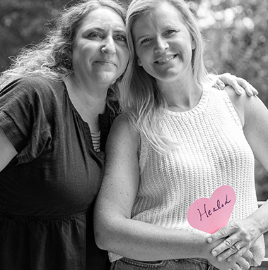 Two friends embrace; one holds a Project Pink heart that reads ‘HEALED’ to celebrate her breast cancer recovery